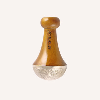 Photograph of the Art of Vedas Kansa Vatki Wand resting on a natural stone surface, highlighting its sleek design and ergonomic handle. The Kansa metal head shines prominently, reflecting its quality and suitability for both foot and body massage.